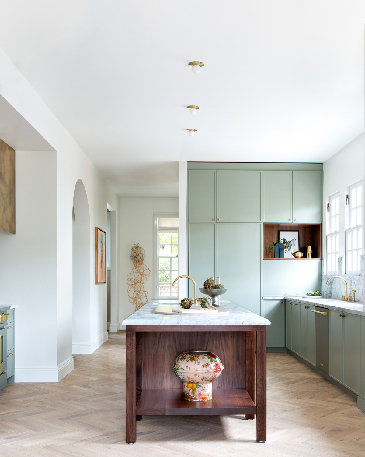 A walnut-toned island sits in the middle of a clean white kitchen with sage cabinetry and minimal finishes. RED Winner.